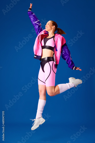 Fototapeta Naklejka Na Ścianę i Meble -  Full-length image of young smiling girl in stylish sportswear cheerfully jumping against blue studio background. Concept of sport, hobby, fashion, youth, human emotions, lifestyle