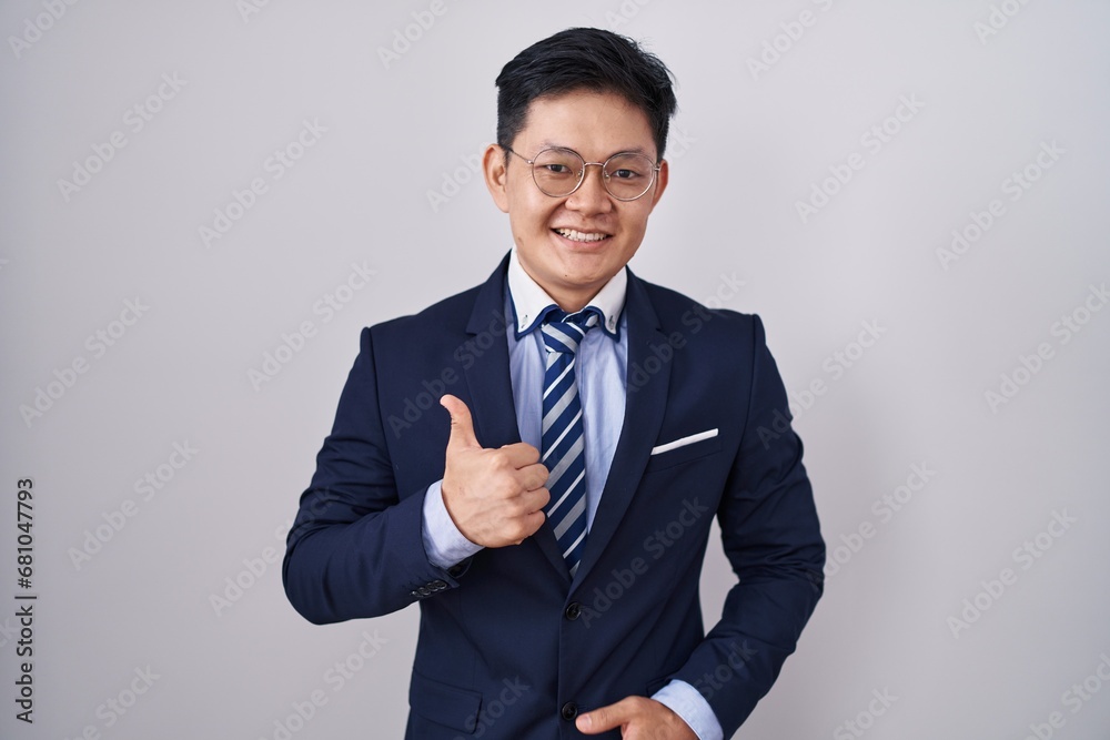 Young asian man wearing business suit and tie doing happy thumbs up gesture with hand. approving expression looking at the camera showing success. Young asian man wearing business suit and tie doing happy thumbs up gesture with hand. approving expression looking at the camera showing success.