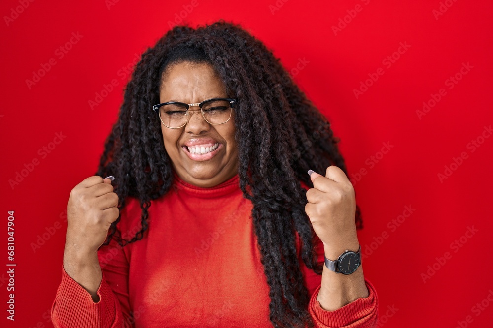 Plus size hispanic woman standing over red background very happy and excited doing winner gesture with arms raised, smiling and screaming for success. celebration concept. Plus size hispanic woman standing over red background very happy and excited doing winner gesture with arms raised, smiling and screaming for success. celebration concept.
