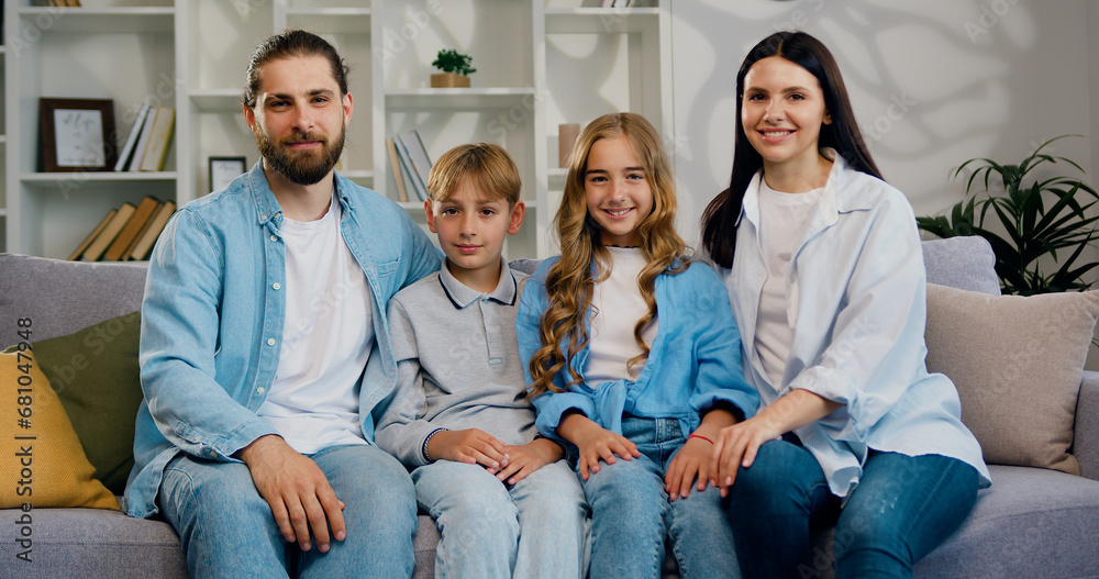 Family portrait of father, mother and two lovely children smiling at camera and hugging in modern living room at home. Good looking caucasian woman and man witch son and daughter sitting on the sofa Family portrait of father, mother and two lovely children smiling at camera and hugging in modern living room at home. Good looking caucasian woman and man witch son and daughter sitting on the sofa
