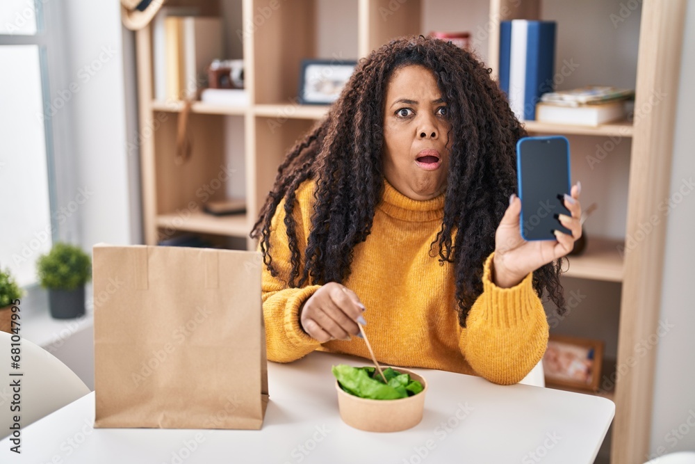 Plus size hispanic woman eating take away salad holding smartphone afraid and shocked with surprise and amazed expression, fear and excited face. Plus size hispanic woman eating take away salad holding smartphone afraid and shocked with surprise and amazed expression, fear and excited face.