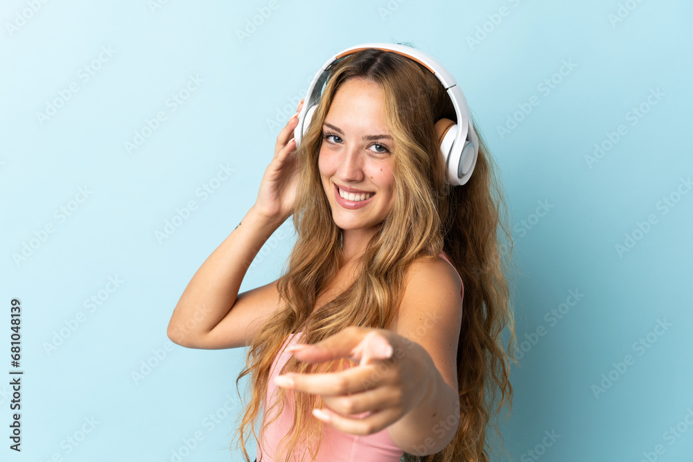 Young blonde woman isolated on blue background listening music and pointing to the front - obrazy, fototapety, plakaty 