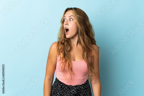 Young blonde woman isolated on blue background doing surprise gesture while looking to the side photo