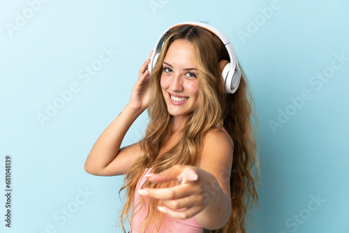 Young blonde woman isolated on blue background listening music and pointing to the front