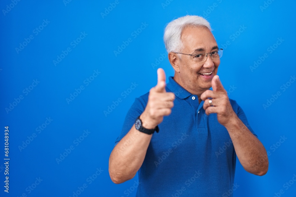 Middle age man with grey hair standing over blue background pointing fingers to camera with happy and funny face. good energy and vibes. Middle age man with grey hair standing over blue background pointing fingers to camera with happy and funny face. good energy and vibes.
