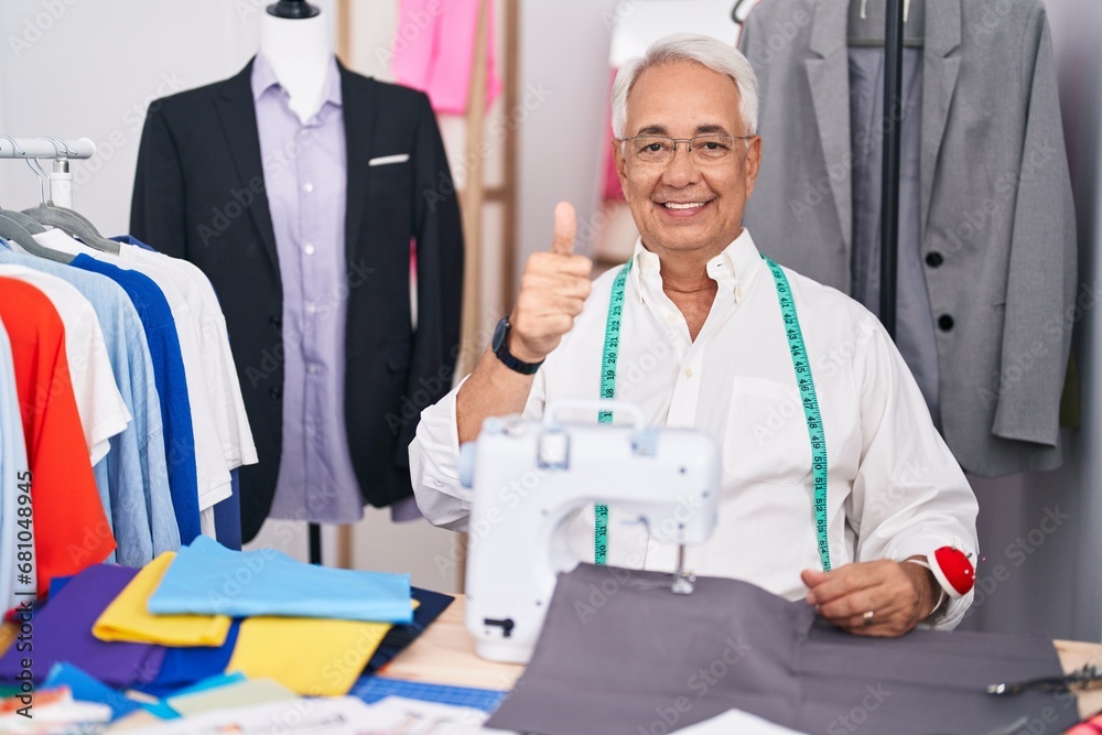 Middle age man with grey hair dressmaker using sewing machine doing happy thumbs up gesture with hand. approving expression looking at the camera showing success. Middle age man with grey hair dressmaker using sewing machine doing happy thumbs up gesture with hand. approving expression looking at the camera showing success.