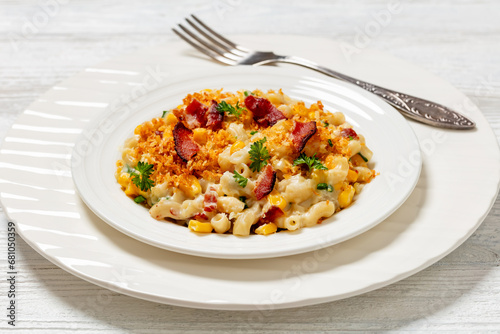 portion of mac and cheese with corn, bacon topped with panko breadcrumbs on white plate on white wooden table, horizontal view from above, close-u