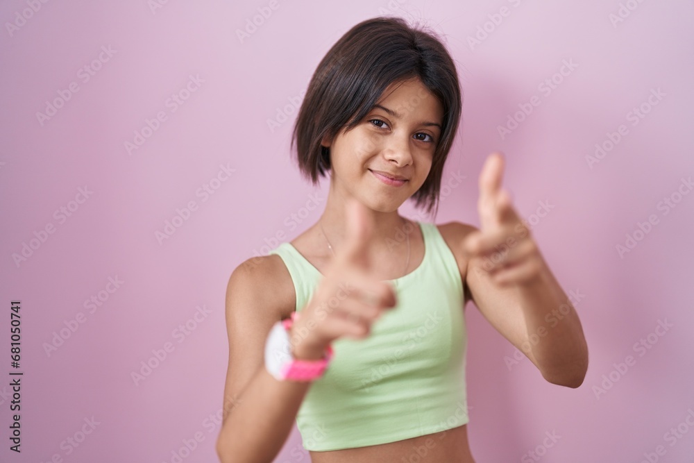 Young girl standing over pink background pointing fingers to camera with happy and funny face. good energy and vibes.