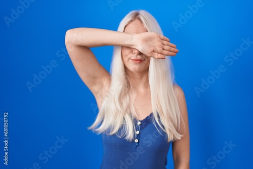 Caucasian woman standing over blue background covering eyes with arm, looking serious and sad. sightless, hiding and rejection concept