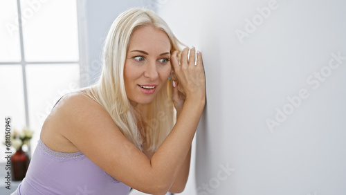 Nosy young blonde woman spied eavesdropping curiosity, glass against wall, listenting in secret at her home's living room, indoor portrait, pyjama clad, beautiful caucasian