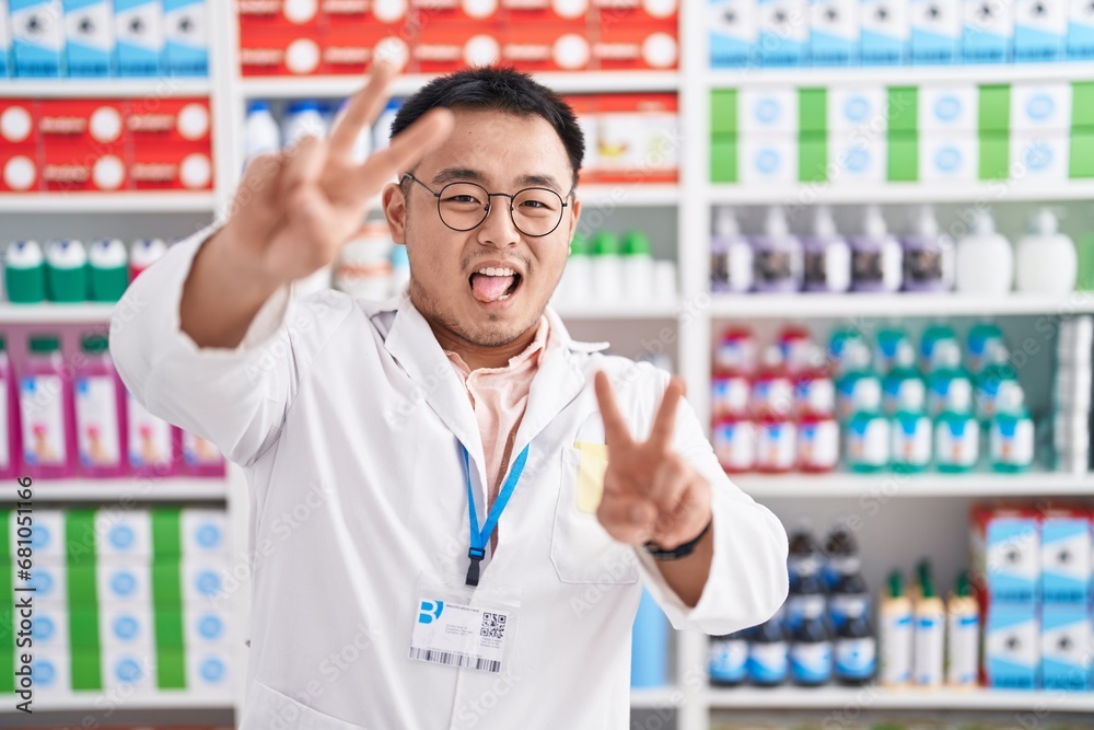 Chinese young man working at pharmacy drugstore smiling with tongue out showing fingers of both hands doing victory sign. number two.