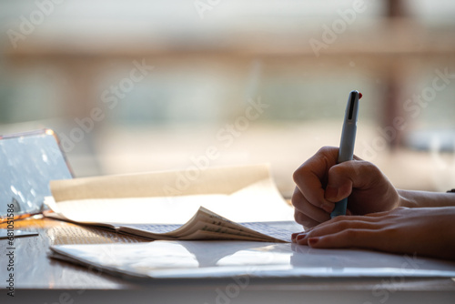 Action of a business person using ballpoint pen to writing on paper sheet to complete the financial report, sitting closed to window with sunlight as background. Selective focus.