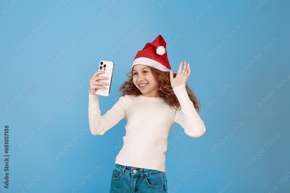 Little African-american girl with Santa hat holding phone and takes selfie on blue background. Little African-american girl with Santa hat holding phone and takes selfie on blue background.