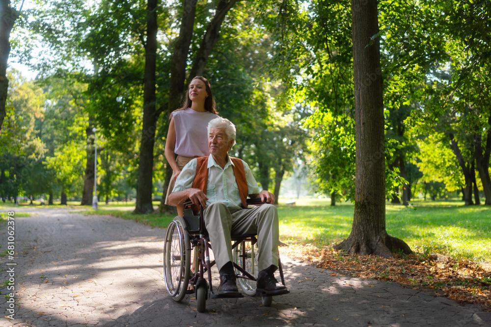 Granddaughter pushing grandfather in wheelchair in city park. Young caregiver spending quality time with lonely senior client in the city park. Granddaughter pushing grandfather in wheelchair in city park. Young caregiver spending quality time with lonely senior client in the city park.