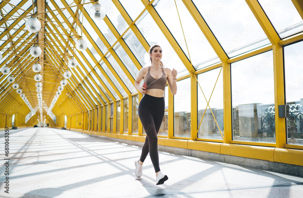 young Caucasian woman in her 20s jogging in a modern yellow pedestrian bridge with glass walls and spherical lamps. She is wearing a brown sports bra, black leggings, and white sneakers young Caucasian woman in her 20s jogging in a modern yellow pedestrian bridge with glass walls and spherical lamps. She is wearing a brown sports bra, black leggings, and white sneakers