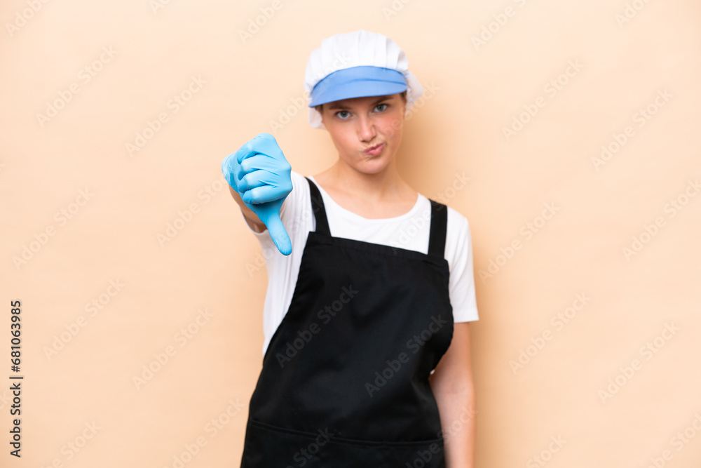 Fishmonger caucasian woman wearing an apron and holding a raw fish isolated on beige background showing thumb down with negative expression Fishmonger caucasian woman wearing an apron and holding a raw fish isolated on beige background showing thumb down with negative expression