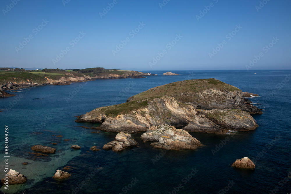 a rocky coastline with a small island covered in greenery, clear blue water, and a clear blue sky
