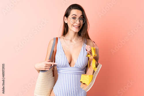Young woman in swimsuit in summer holidays holding a beach bag and summer sandals over isolated pink background with surprise facial expression