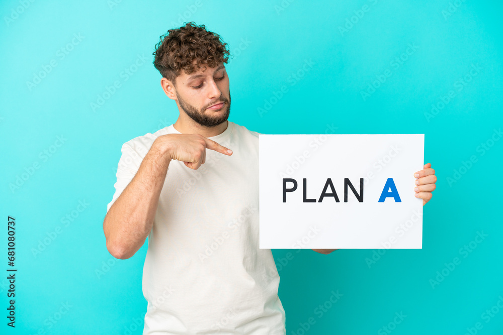 Young handsome caucasian man isolated on blue background holding a placard with the message PLAN A and pointing it Young handsome caucasian man isolated on blue background holding a placard with the message PLAN A and pointing it
