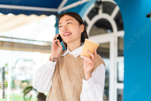 Young Chinese woman at outdoors using mobile phone and holding a coffee with happy expression photo