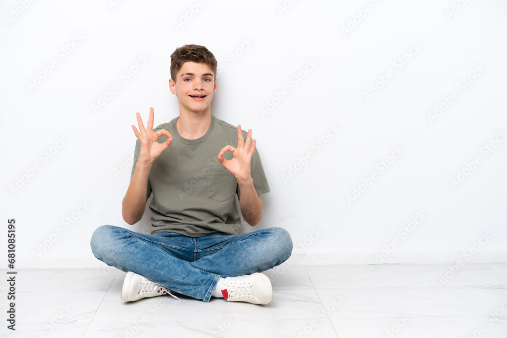 Teenager Russian man sitting on the floor isolated on white background showing an ok sign with fingers Teenager Russian man sitting on the floor isolated on white background showing an ok sign with fingers