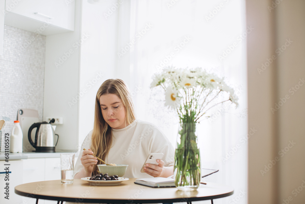 The blonde girl has breakfast while holding the phone in her hands.