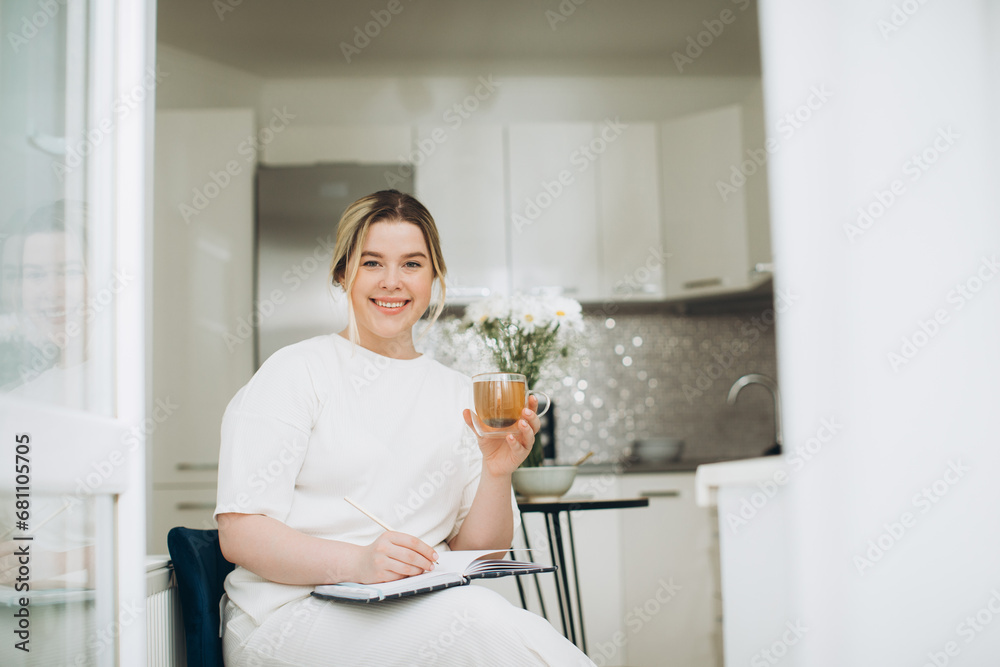 Smiling young woman drinking tea and writing in her diary or creative journal in the morning in the kitchen. Smiling young woman drinking tea and writing in her diary or creative journal in the morning in the kitchen.