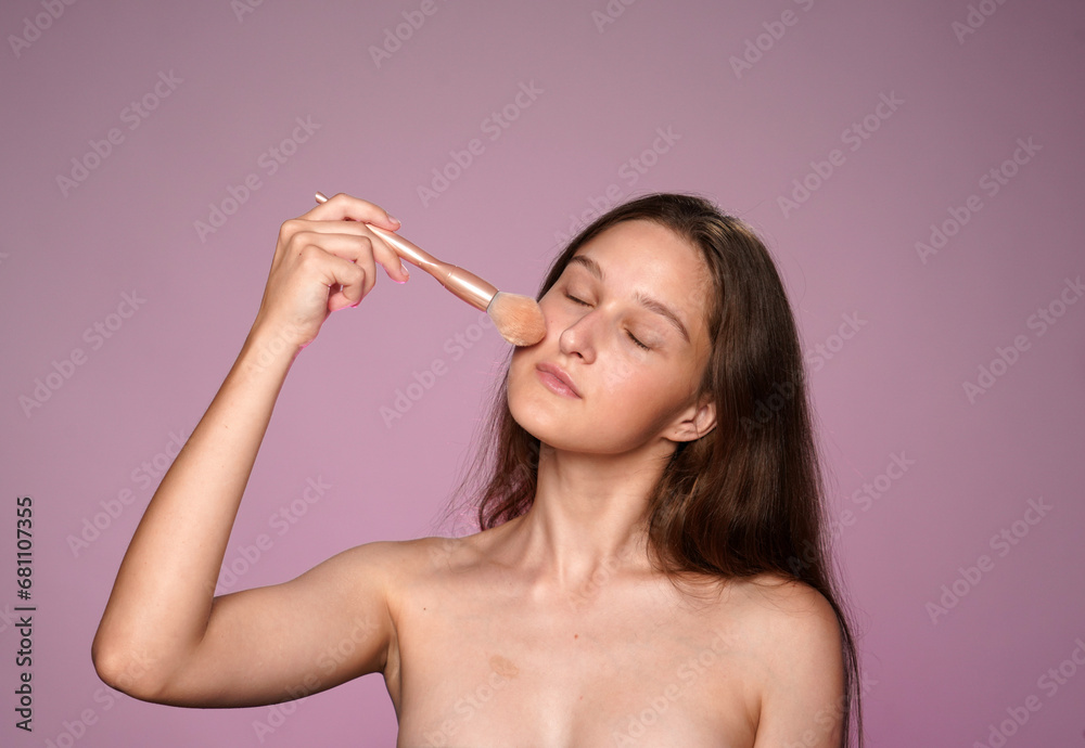 Obraz premium Woman applying makeup using a makeup brush. Seminude woman in studio on pink background in pink neon light close up. Contouring. Concept of beauty, cosmetology, skin care.