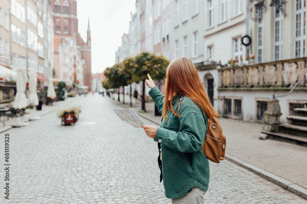 Attractive young female tourist is exploring city. Redhead girl with backpack pointing finger and holding a paper map on city street in Gdansk. Traveling Europe in autumn. - obrazy, fototapety, plakaty 