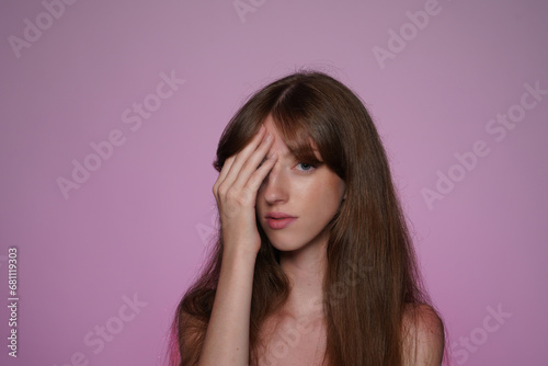 Seminude woman covered her eye with her hand. Longhaired woman in studio on pink background in pink neon light. Natural female beauty. Cosmetic line for eye contour care.