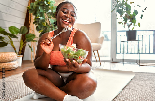 Healthy diet and sport. Beautiful plump woman in sport clothes eating vegetable salad from glass bowl while sitting on floor. Black young female following slimming and exercising program at home. photo
