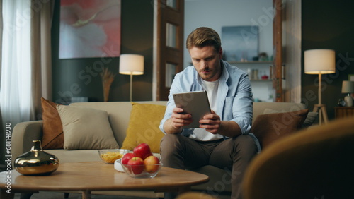 Calm man browsing tab device at home. Serious guy sitting sofa scrolling tablet