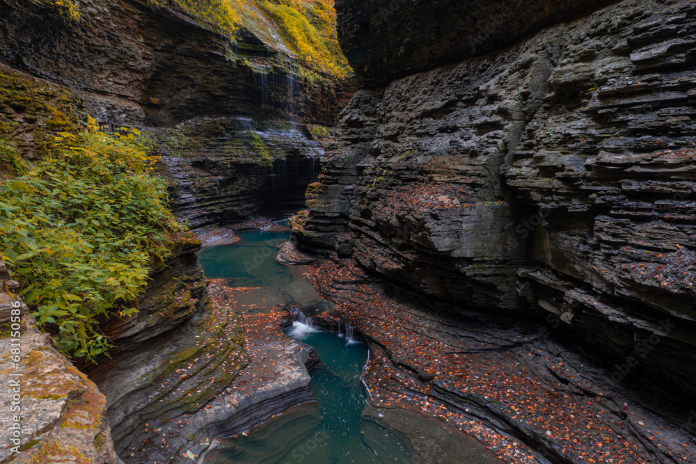 Waterfall in deep limestone gorge surrounded by forest trees during fall month. Water flows smooth over rocky cliffs into deep pool. Long exposure, wide angle shot. Watkins Glen State Park, NY, USA.