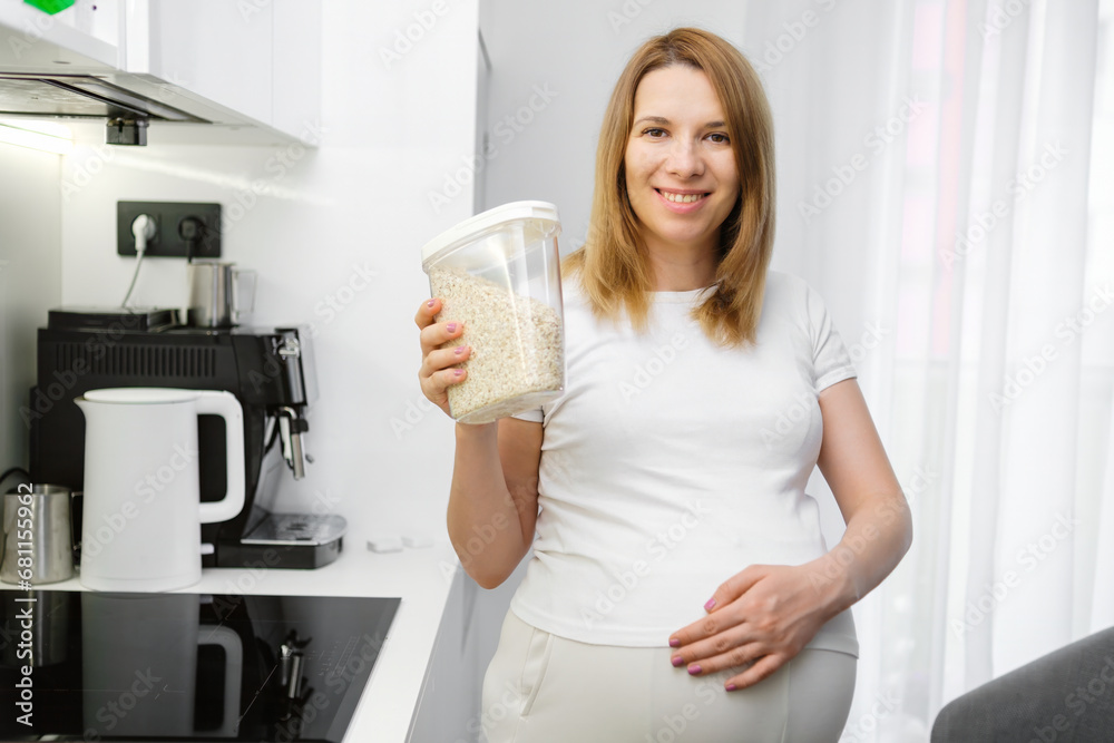 Beautiful pregnant woman holding a container with oats in the kitchen. Diet and vitamins during pregnancy. Beautiful pregnant woman holding a container with oats in the kitchen. Diet and vitamins during pregnancy.