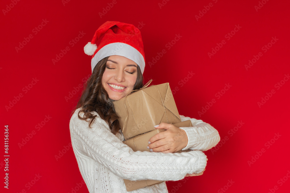 a young pretty brunette woman in a Santa Claus hat received gifts. She hugs gifts wrapped in kraft paper, smiles, closes her eyes with happiness. Isolated on a red background. a young pretty brunette woman in a Santa Claus hat received gifts. She hugs gifts wrapped in kraft paper, smiles, closes her eyes with happiness. Isolated on a red background.