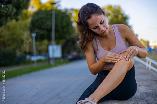 Woman with knee pain in a park. Female athlete suffering form running knee or kneecap injury during outdoor workout. Woman sitting on ground and holding knee