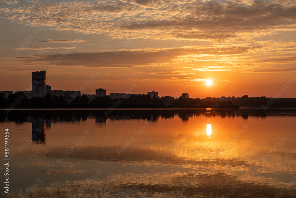 Scenic sunrise sun shining on lake with reflection on calm mirror water surface. Colorful morning by the river in city park with multistory buildings Scenic sunrise sun shining on lake with reflection on calm mirror water surface. Colorful morning by the river in city park with multistory buildings