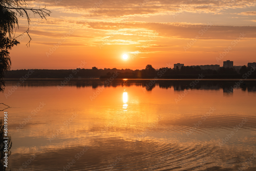 Scenic sunrise sun shining on lake with clouds reflecting in calm water surface. Colorful morning by the river in city park with multistory buildings Scenic sunrise sun shining on lake with clouds reflecting in calm water surface. Colorful morning by the river in city park with multistory buildings