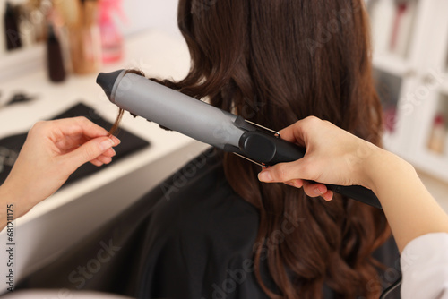 Hairdresser working with client using curling hair iron in salon, closeup