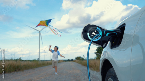 Focus EV car recharging display battery status hologram, charging station using eco-friendly energy wind turbine generator with happy young boy running and playing with kite in background. Peruse
