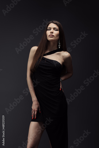 A stunning model poses in an asymmetrical black dress, her red nail polish complementing her striking appearance against a dark backdrop