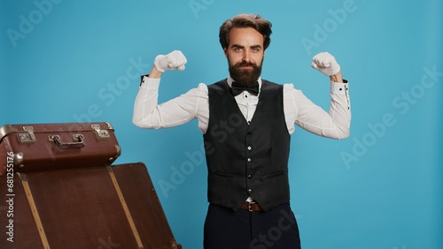 Modern bellboy flexing muscles in studio, showing off strength underneath his formal uniform attire. Hotel porter doorman feeling powerful on camera, showing determination in hospitality industry. photo