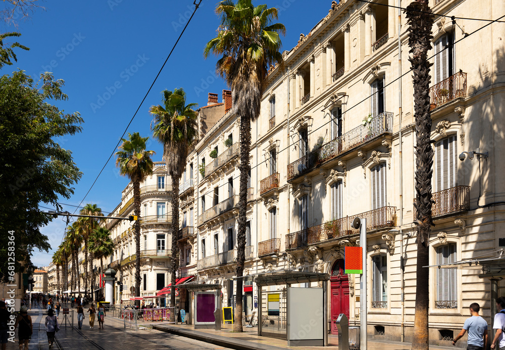 View of vibrant cobbled street with tram tracks, typical architecture and tall palm trees in French city of Montpellier on sunny summer day .