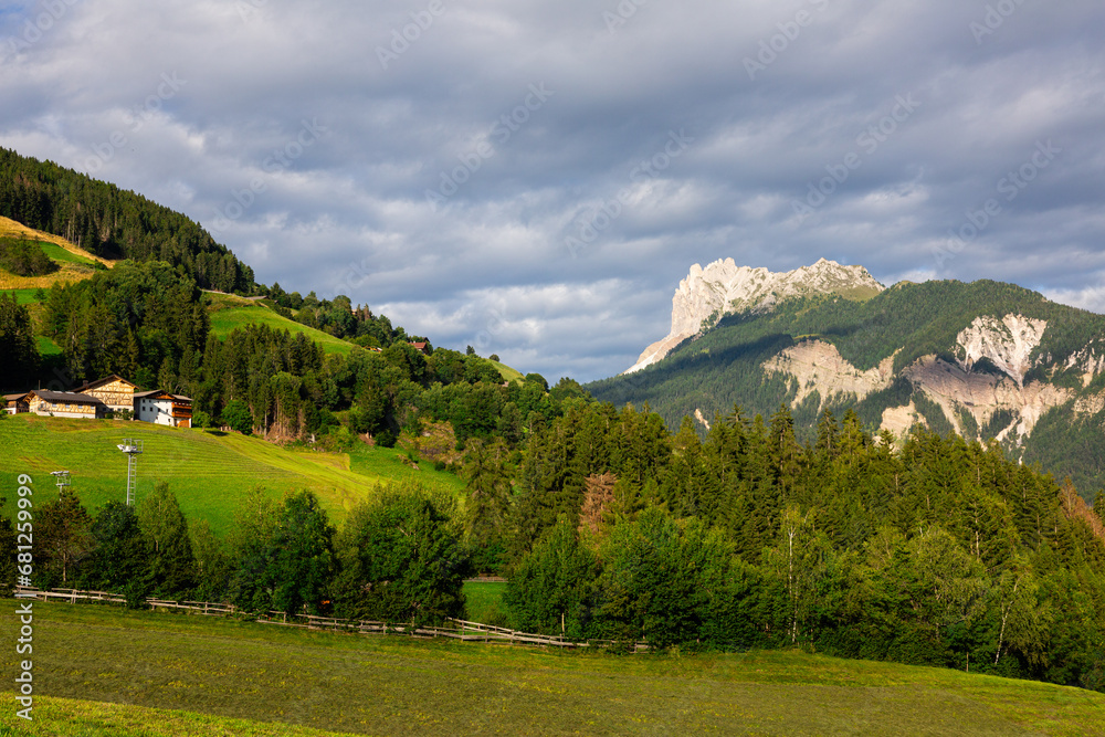 Scenic view of vibrant green alpine valley of Val di Funes with lush meadows, forested hills and cozy settlements surrounded by rugged rocky range of Italian Dolomites on sunny summer day