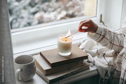 Kids hand in checkered pyjamas with burning match lighting candle on windowsill. Autumn still life with cup of coffee, oldbooks. Autumn home decor. Blurred garden background. Cozy fall, winter mood. photo