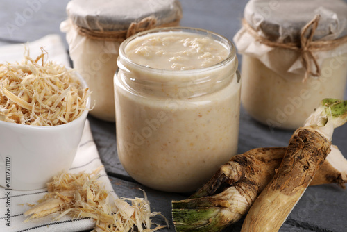 Leinwand Poster Spicy horseradish sauce in jars and roots on grey wooden table, closeup