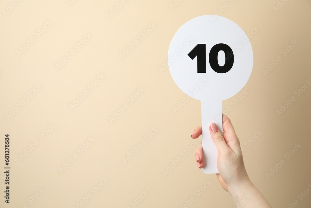 Woman holding auction paddle with number 10 on beige background, closeup. Space for text