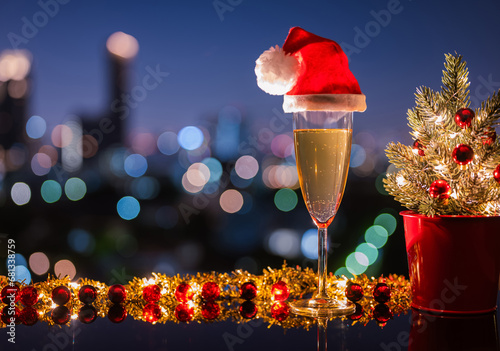 A glass of white wine that have santa claus hat on it with Christmas ornament decoration put on table with city bokeh light background. Christmas dining concept. photo