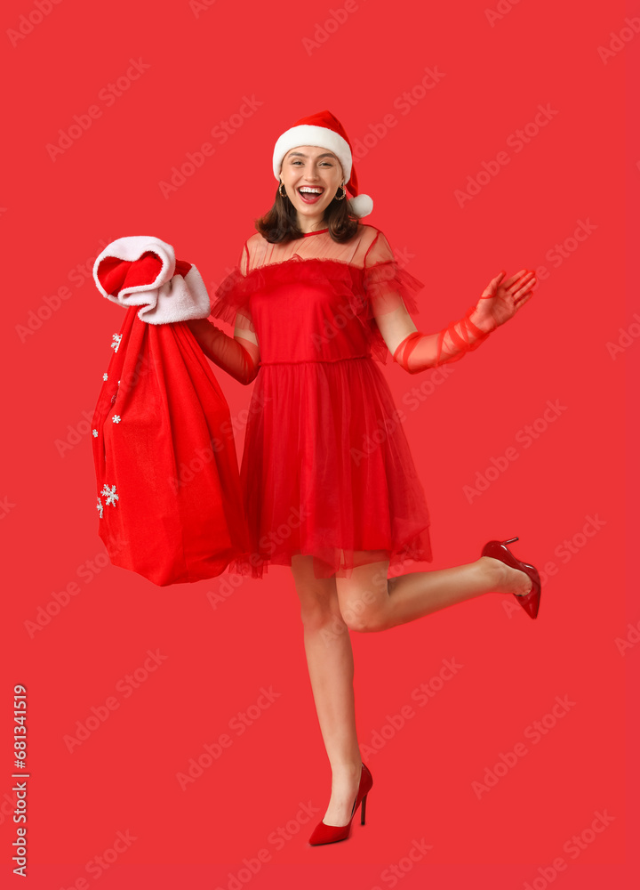 Happy young woman in Santa hat holding bag with Christmas gifts on red background