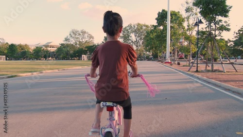 Rear view showing back of 7 years old sporty Asian kid girl are riding bicycle for active outdoor living. Her winning smile and energetic spirit infuse the scene with joy and happiness.